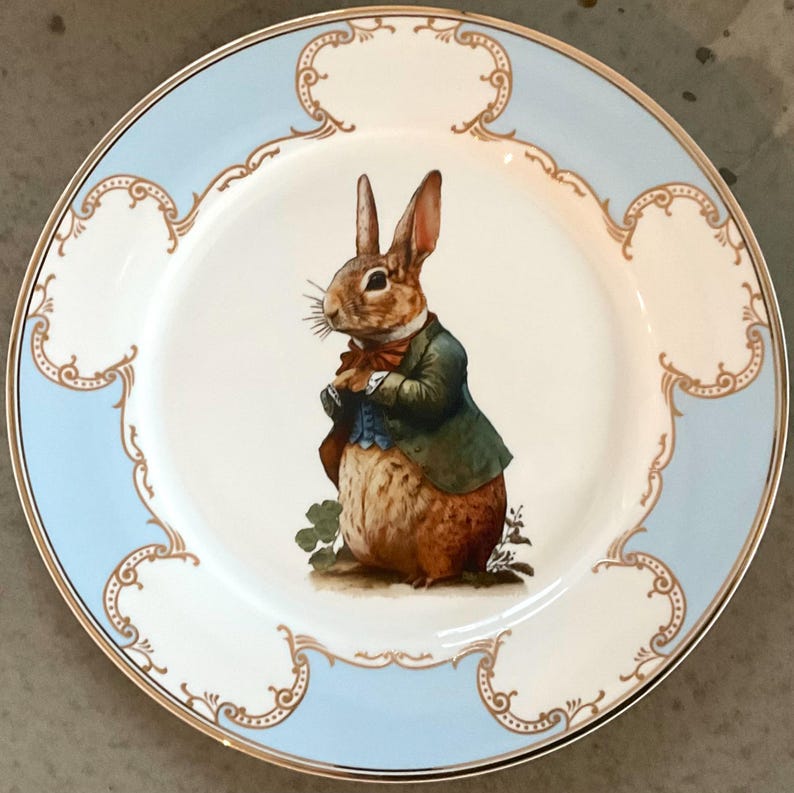 Peut inclure: Une assiette en c&eacute;ramique blanche avec un bord bleu et une bordure dor&eacute;e pr&eacute;sente une illustration peinte &agrave; la main d'un lapin brun portant une veste verte et un n&oelig;ud papillon bleu. Le lapin est debout sur une touffe d'herbe verte avec du tr&egrave;fle.