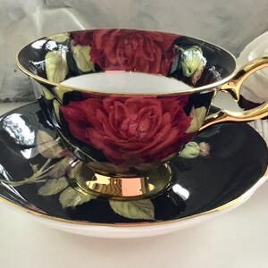 Puede incluir: Una taza de t&eacute; y un platillo negros con un borde dorado y un patr&oacute;n de rosas rojas. La taza tiene un asa dorada.