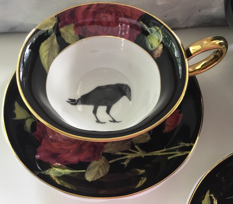 Puede incluir: Un juego de taza y platillo de t&eacute; negro y dorado con una silueta de cuervo negro en el interior de la taza y un dise&ntilde;o de rosa roja en el exterior.