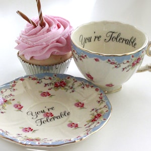 May include: A white teacup and saucer with a blue and floral pattern. The teacup and saucer both have the text "You're Tolerable" printed on them. A pink cupcake with chocolate sprinkles is in the background.