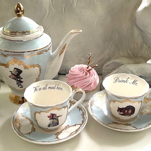 Puede incluir: Un juego de t&eacute; de color azul y dorado con tem&aacute;tica de Alicia en el Pa&iacute;s de las Maravillas, que incluye una tetera, un azucarero, una jarra de crema y dos tazas y platillos. Las tazas muestran las frases "We're all mad here." y "Drink Me."