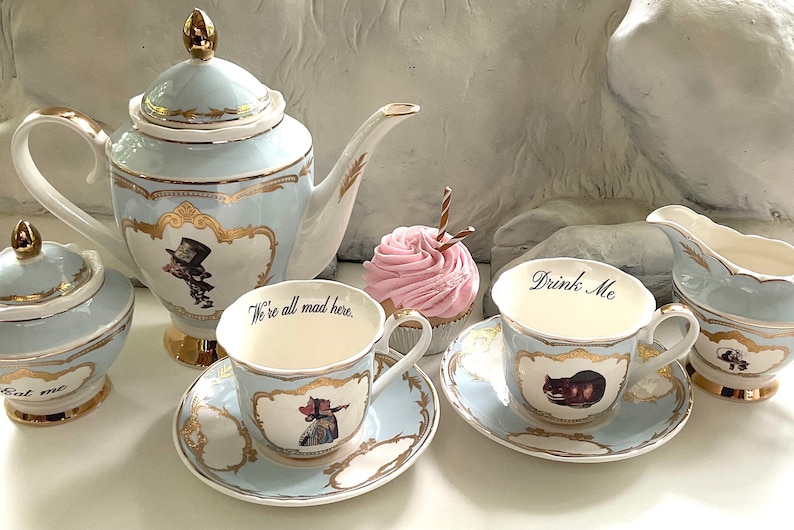Puede incluir: Un juego de t&eacute; de color azul y dorado con tem&aacute;tica de Alicia en el Pa&iacute;s de las Maravillas, que incluye una tetera, un azucarero, una jarra de crema y dos tazas y platillos. Las tazas muestran las frases "We're all mad here." y "Drink Me."