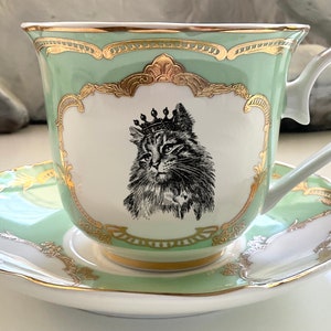 K&ouml;nnte beinhalten: Ein gr&uuml;nes Teetassen- und Untertassen-Set mit goldener Verzierung und einer Schwarzwei&szlig;illustration einer Katze mit Krone.