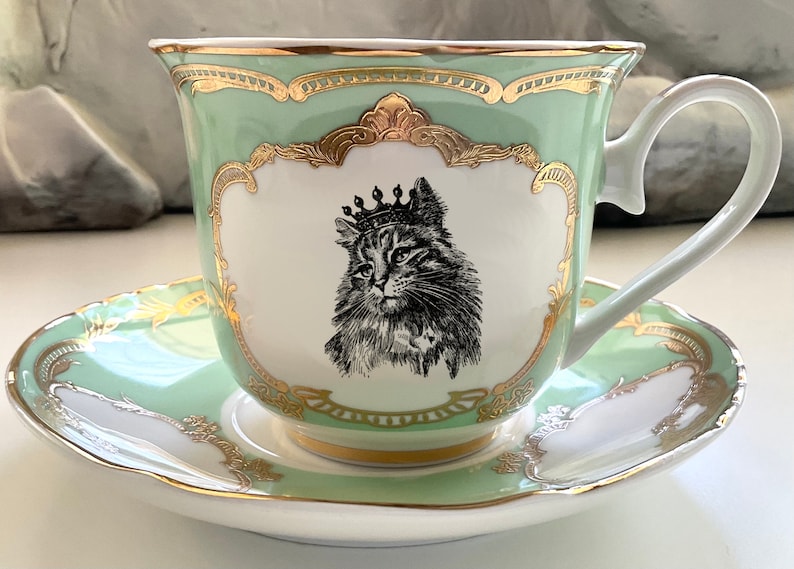 K&ouml;nnte beinhalten: Ein gr&uuml;nes Teetassen- und Untertassen-Set mit goldener Verzierung und einer Schwarzwei&szlig;illustration einer Katze mit Krone.