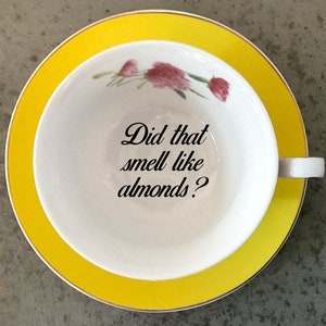 May include: A white teacup with a yellow saucer and a floral design. The teacup has the text "Did that smell like almonds?" printed on the inside.
