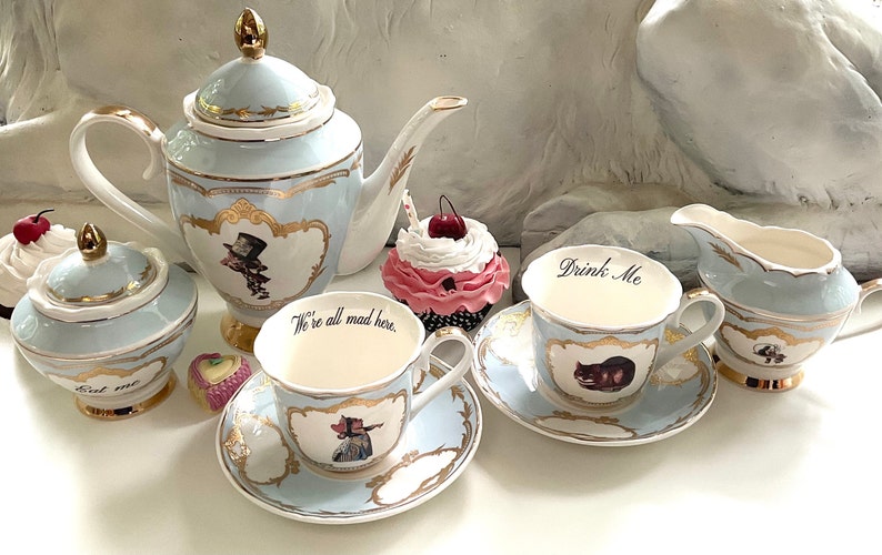 Puede incluir: Un juego de t&eacute; de color azul y dorado con tem&aacute;tica de Alicia en el Pa&iacute;s de las Maravillas, que incluye una tetera, un azucarero, una jarra de crema y dos tazas de t&eacute; con sus platillos. Las tazas de t&eacute; tienen las frases "We're all mad here." y "Drink Me" en ellas.