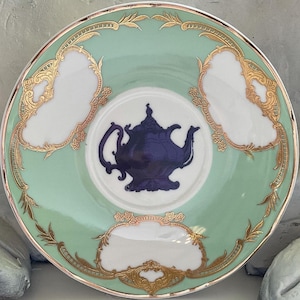 Puede incluir: Un platillo de porcelana verde y blanco con ribetes dorados y una silueta azul de una tetera en el centro.