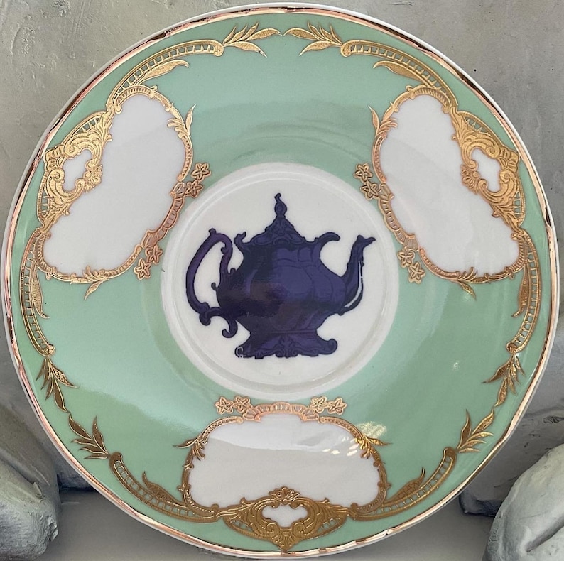 Puede incluir: Un platillo de porcelana verde y blanco con ribetes dorados y una silueta azul de una tetera en el centro.