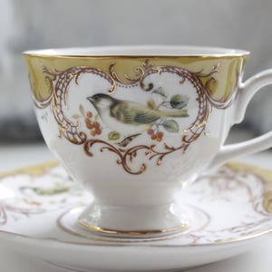 May include: A white teacup and saucer with a gold rim and intricate floral and bird design. The cup has a delicate handle and the saucer has a yellow rim.