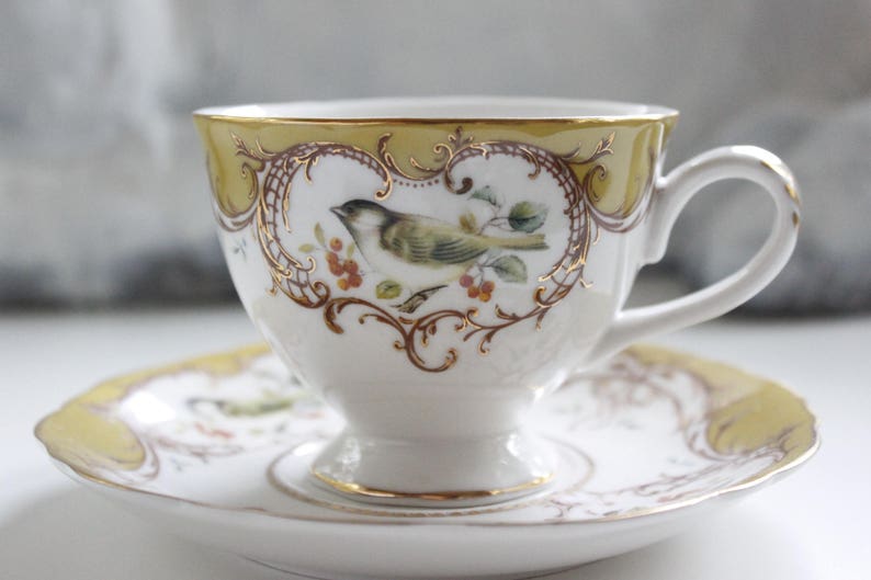 May include: A white teacup and saucer with a gold rim and intricate floral and bird design. The cup has a delicate handle and the saucer has a yellow rim.