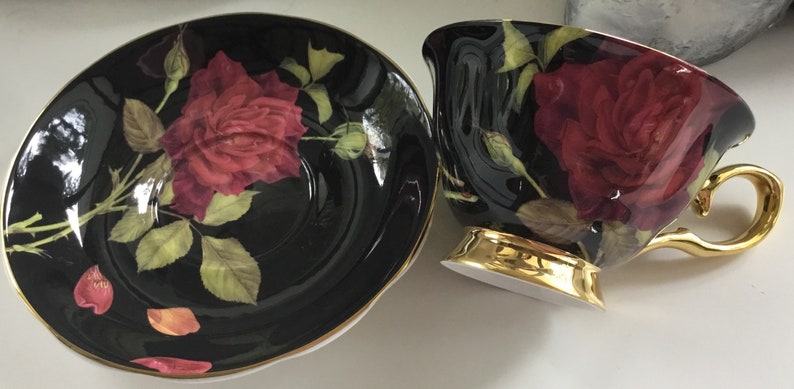 Puede incluir: Un juego de taza y platillo de t&eacute; negro con borde dorado. El juego presenta un dise&ntilde;o de rosa roja sobre fondo negro.