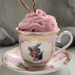 Puede incluir: Una taza de t&eacute; y un platillo rosas con ribetes dorados presentan una ilustraci&oacute;n caprichosa del Conejo Blanco de Alicia en el Pa&iacute;s de las Maravillas sosteniendo una carta de juego. La taza de t&eacute; est&aacute; llena de crema batida rosa y rematada con dos palitos de chocolate.