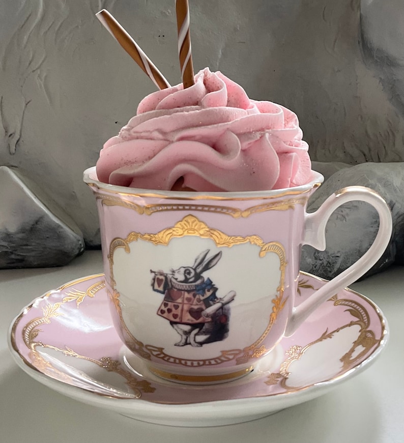 Puede incluir: Una taza de t&eacute; y un platillo rosas con ribetes dorados presentan una ilustraci&oacute;n caprichosa del Conejo Blanco de Alicia en el Pa&iacute;s de las Maravillas sosteniendo una carta de juego. La taza de t&eacute; est&aacute; llena de crema batida rosa y rematada con dos palitos de chocolate.