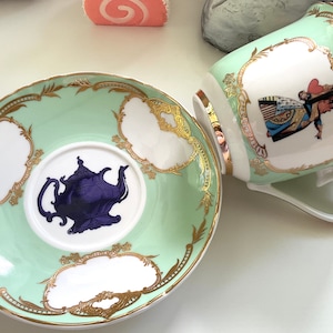 Puede incluir: Un juego de taza y platillo de t&eacute; verde y dorado con un dise&ntilde;o de tetera morada en el platillo y una ilustraci&oacute;n de la Reina de Corazones en la taza.