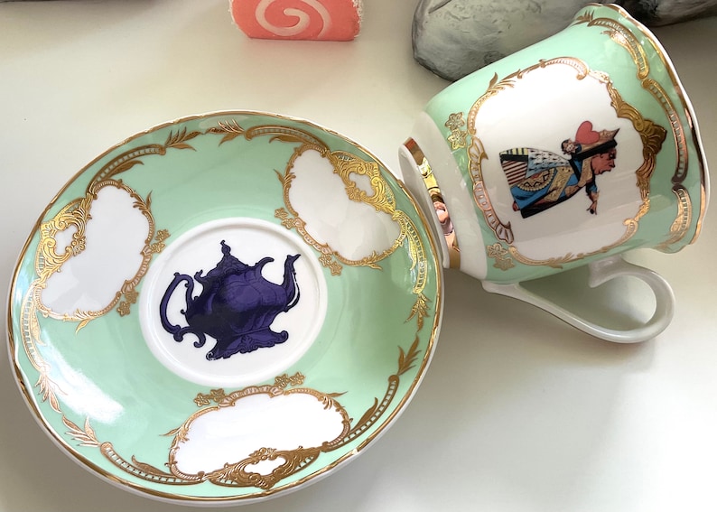 Puede incluir: Un juego de taza y platillo de t&eacute; verde y dorado con un dise&ntilde;o de tetera morada en el platillo y una ilustraci&oacute;n de la Reina de Corazones en la taza.