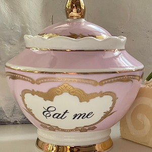 Puede incluir: Un tarro de cer&aacute;mica rosa y dorado con tapa. El tarro tiene un borde dorado y un dise&ntilde;o dorado. El tarro tiene las palabras "Eat me" escritas en &eacute;l.