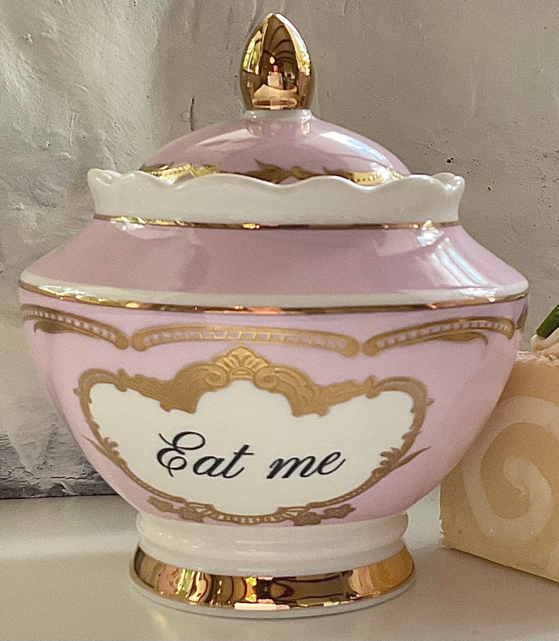 Puede incluir: Un tarro de cer&aacute;mica rosa y dorado con tapa. El tarro tiene un borde dorado y un dise&ntilde;o dorado. El tarro tiene las palabras "Eat me" escritas en &eacute;l.