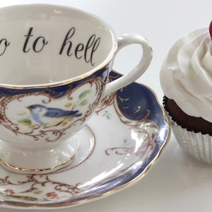 May include: A white teacup and saucer with blue and gold trim and a floral design. The teacup has the words "Go to hell" written on the inside. A red and white cupcake with white frosting is next to the teacup.