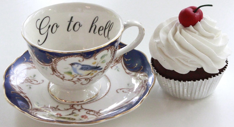 May include: A white teacup and saucer with blue and gold trim and a floral design. The teacup has the words "Go to hell" written on the inside. A red and white cupcake with white frosting is next to the teacup.