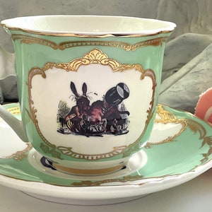 Puede incluir: Una taza de t&eacute; verde y un platillo con ribete dorado y una ilustraci&oacute;n de un conejo blanco. El platillo tiene un rollo de pastel rosa y blanco encima.