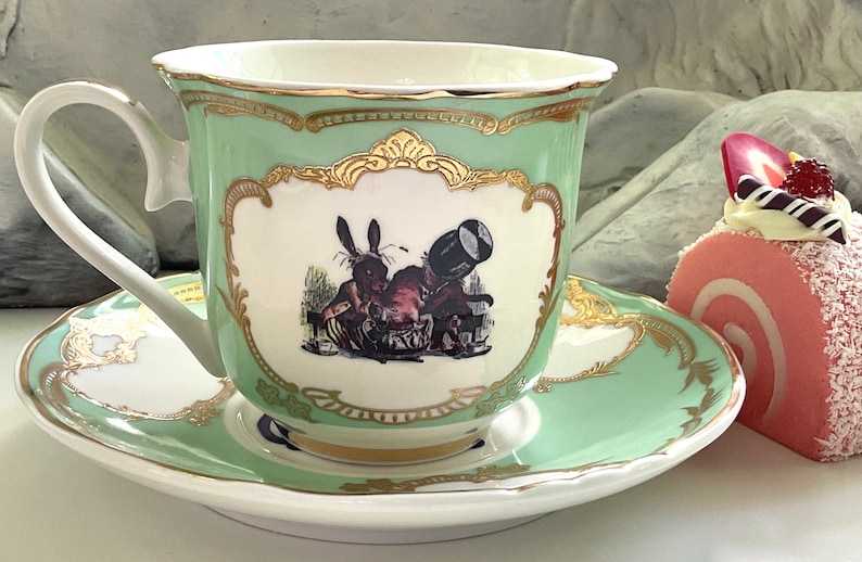 Puede incluir: Una taza de t&eacute; verde y un platillo con ribete dorado y una ilustraci&oacute;n de un conejo blanco. El platillo tiene un rollo de pastel rosa y blanco encima.