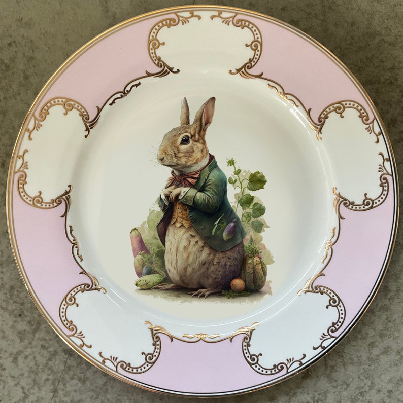 Peut inclure: Une assiette en c&eacute;ramique blanche avec un bord rose et une bordure dor&eacute;e pr&eacute;sente une illustration fantaisiste d'un lapin portant un gilet et tenant une fleur. L'assiette est parfaite pour servir un repas sp&eacute;cial ou ajouter une touche de charme &agrave; votre d&eacute;coration de cuisine.