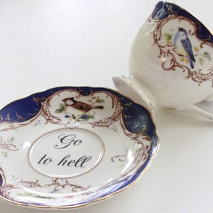 May include: A blue and gold teacup and saucer set with a floral design and a bird. The saucer has the text "Go to hell" written on it.