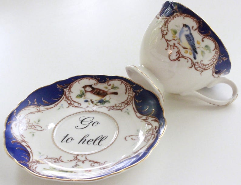 May include: A blue and gold teacup and saucer set with a floral design and a bird. The saucer has the text "Go to hell" written on it.
