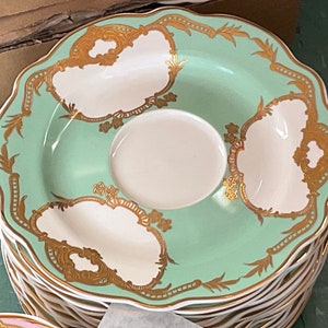 K&ouml;nnte beinhalten: Ein Stapel gr&uuml;ner und wei&szlig;er Untertassen aus Keramik mit goldenen Verzierungen und einem zarten Blumenmuster. Die Untertassen haben einen gewellten Rand und eignen sich perfekt zum Servieren von Tee oder Kaffee.