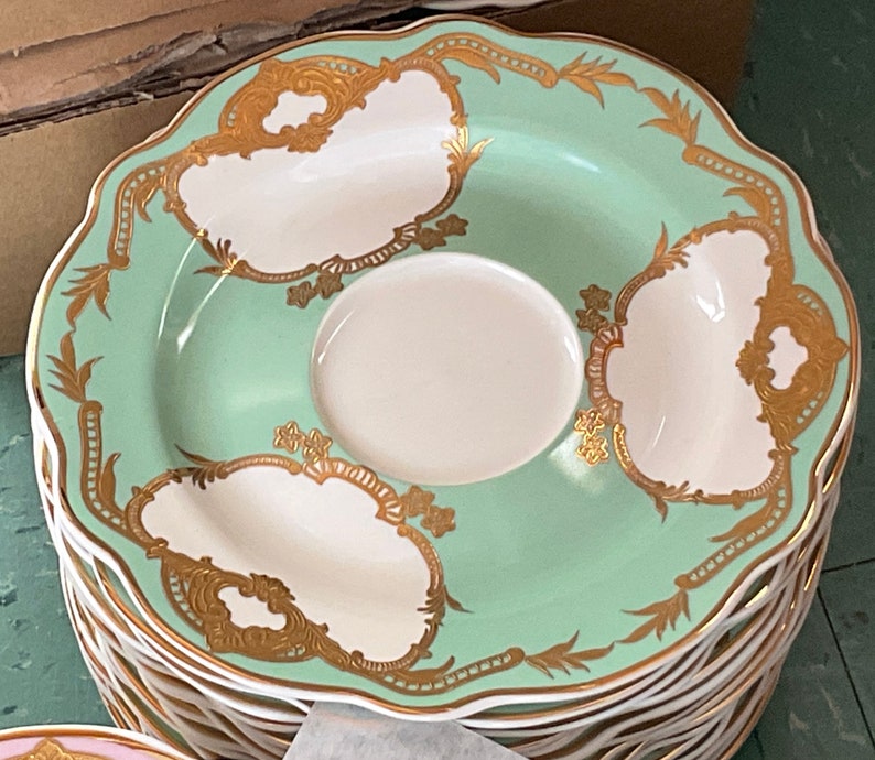 K&ouml;nnte beinhalten: Ein Stapel gr&uuml;ner und wei&szlig;er Untertassen aus Keramik mit goldenen Verzierungen und einem zarten Blumenmuster. Die Untertassen haben einen gewellten Rand und eignen sich perfekt zum Servieren von Tee oder Kaffee.