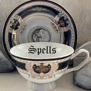 May include: A white teacup and saucer with a black and gold design. The teacup has the word "Spells" written in black script on the inside. The saucer has a black and gold design with a crescent moon and stars.