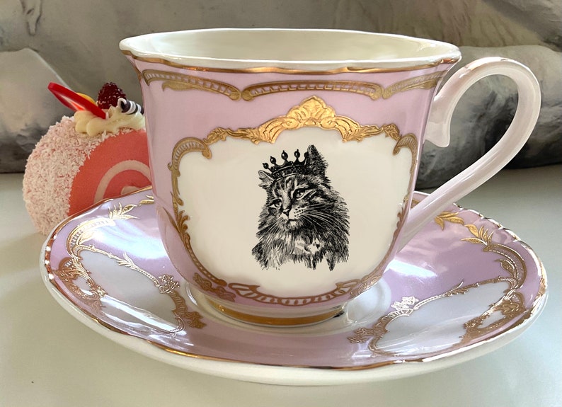K&ouml;nnte beinhalten: Eine rosa Teetasse und Untertasse mit goldenen Verzierungen und einer Schwarzwei&szlig;illustration einer Katze mit Krone. Die Teetasse hat einen gewellten Rand und die Untertasse ein zartes Blumenmuster.