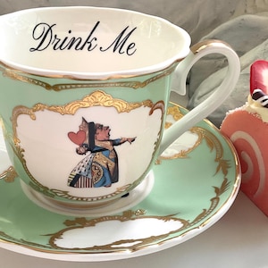 Puede incluir: Una taza de t&eacute; verde y un platillo con ribetes dorados y una representaci&oacute;n de la Reina de Corazones de Alicia en el Pa&iacute;s de las Maravillas. El texto "Drink Me" est&aacute; escrito en la taza. Un rollo de pastel rosa y blanco con una fresa en la parte superior est&aacute; al lado de la taza de t&eacute;.
