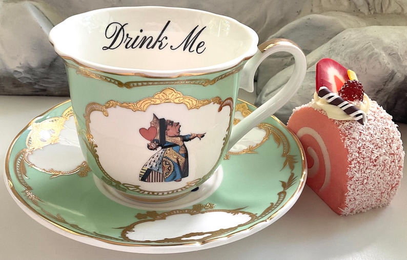 Puede incluir: Una taza de t&eacute; verde y un platillo con ribetes dorados y una representaci&oacute;n de la Reina de Corazones de Alicia en el Pa&iacute;s de las Maravillas. El texto "Drink Me" est&aacute; escrito en la taza. Un rollo de pastel rosa y blanco con una fresa en la parte superior est&aacute; al lado de la taza de t&eacute;.