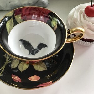 Puede incluir: Una taza de t&eacute; y un platillo negros con un borde dorado. La taza de t&eacute; presenta un murci&eacute;lago negro volando sobre un fondo blanco. El platillo tiene un fondo negro con un dise&ntilde;o floral en tonos rojos, verdes y dorados.