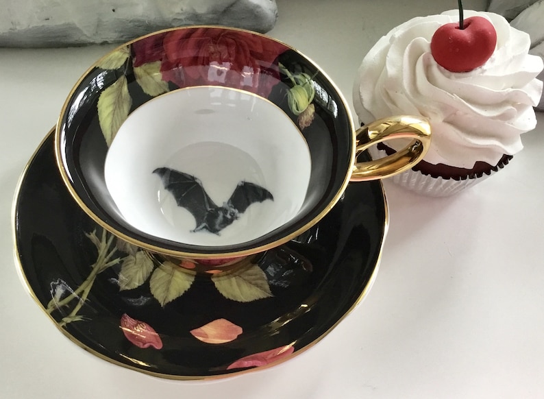 Puede incluir: Una taza de t&eacute; y un platillo negros con un borde dorado. La taza de t&eacute; presenta un murci&eacute;lago negro volando sobre un fondo blanco. El platillo tiene un fondo negro con un dise&ntilde;o floral en tonos rojos, verdes y dorados.