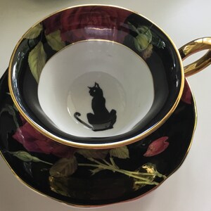 Puede incluir: Una taza de t&eacute; y un platillo negros y dorados con una silueta de gato negro dentro de la taza. El platillo presenta un dise&ntilde;o floral con rosas rojas y hojas verdes sobre un fondo negro.
