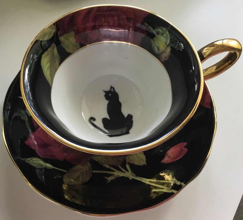 Puede incluir: Una taza de t&eacute; y un platillo negros y dorados con una silueta de gato negro dentro de la taza. El platillo presenta un dise&ntilde;o floral con rosas rojas y hojas verdes sobre un fondo negro.