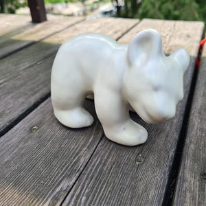 Op de afbeelding: Een kleine, crèmekleurige keramische berenfiguur. De beer wordt afgebeeld in een staande positie met het hoofd iets naar beneden. Het beeldje heeft een gladde, glanzende afwerking en staat op een verweerd houten oppervlak.
