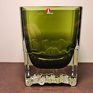 Può includere: Un vaso quadrato in vetro verde oliva con una base trasparente e strutturata. Il vaso presenta un motivo ondulato al centro e un adesivo rosso e bianco sul bordo superiore. Il vaso è appoggiato su una superficie marrone.