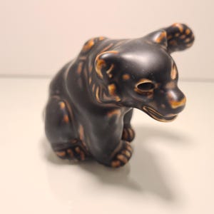 Può includere: Statuetta di orso in ceramica nera con dettagli marroni su orecchie, zampe e muso. L'orso è raffigurato in posizione seduta e ha una finitura lucida. Lo sfondo è di colore neutro.