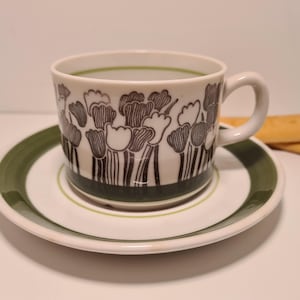 May include: A white ceramic teacup and saucer set with a floral design in black and white. The cup has a handle and sits on a matching saucer with a green border. Two golden-brown cookies are placed next to the saucer.