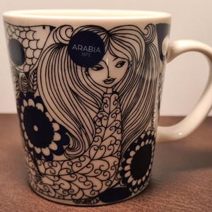 Peut inclure: Tasse en céramique blanche avec une illustration bleu marine d'une femme aux longs cheveux et motifs floraux. La tasse a une anse incurvée et le texte "ARABIA 1873" est visible.