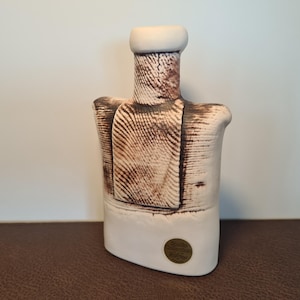 Può includere: Una scultura in ceramica con un design strutturato marrone e bianco. La scultura ha un collo cilindrico e una base rettangolare. Un piccolo adesivo rotondo color oro è sulla base.