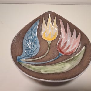May include: A brown ceramic decorative dish with a floral design. The dish features a yellow tulip, a pink tulip, and a blue leaf. The tulips and leaf are raised from the surface of the dish.