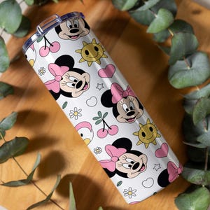 Pu&ograve; includere: Un bicchiere cilindrico bianco con un motivo ripetuto di volti di Minnie con fiocchi rosa, soli gialli sorridenti, ciliegie rosa, piccoli fiori bianchi e cuori rosa, dotato di un coperchio in plastica trasparente.