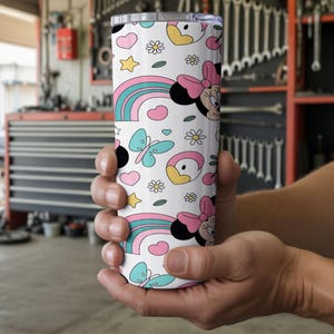 Pu&ograve; includere: Un bicchiere cilindrico bianco con un motivo ripetuto di teste di Minnie con fiocchi rosa, arcobaleni colorati, farfalle azzurre, piccole margherite, stelle gialle e cuori rosa, dotato di un coperchio trasparente.