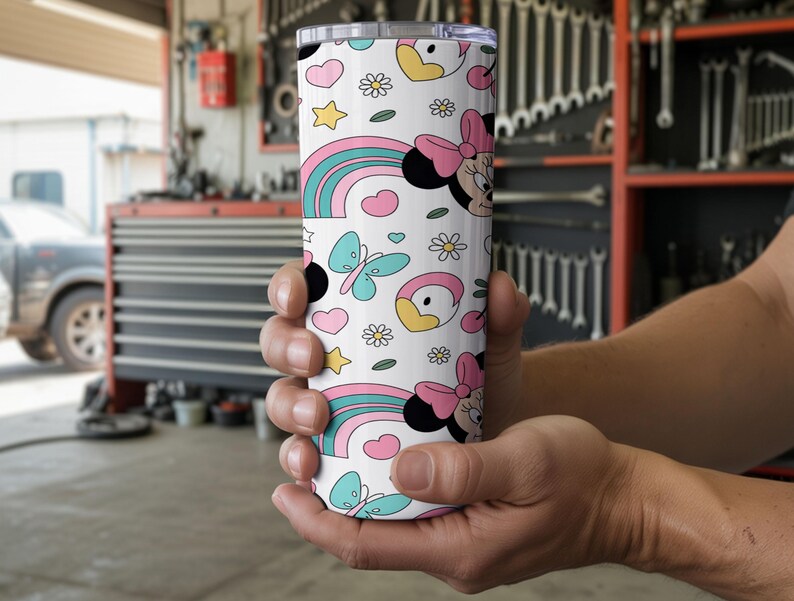 Pu&ograve; includere: Un bicchiere cilindrico bianco con un motivo ripetuto di teste di Minnie con fiocchi rosa, arcobaleni colorati, farfalle azzurre, piccole margherite, stelle gialle e cuori rosa, dotato di un coperchio trasparente.