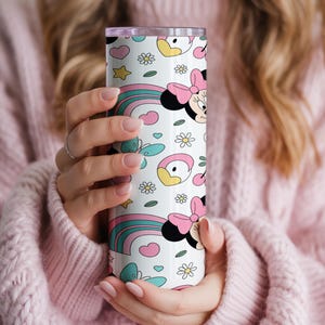 Pu&ograve; includere: Un bicchiere cilindrico in acciaio inossidabile con sfondo bianco e un motivo ripetuto di teste di Minnie Mouse, fiocchi rosa, arcobaleni colorati, farfalle color ottanio, stelle gialle, piccoli cuori e margherite bianche.