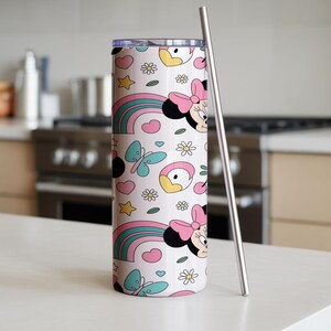 Pu&ograve; includere: Un bicchiere cilindrico con coperchio trasparente e cannuccia in metallo, caratterizzato da un motivo ripetuto su sfondo rosa chiaro. Il design include teste di topo con fiocchi rosa, arcobaleni colorati, farfalle azzurre e fiori bianchi.